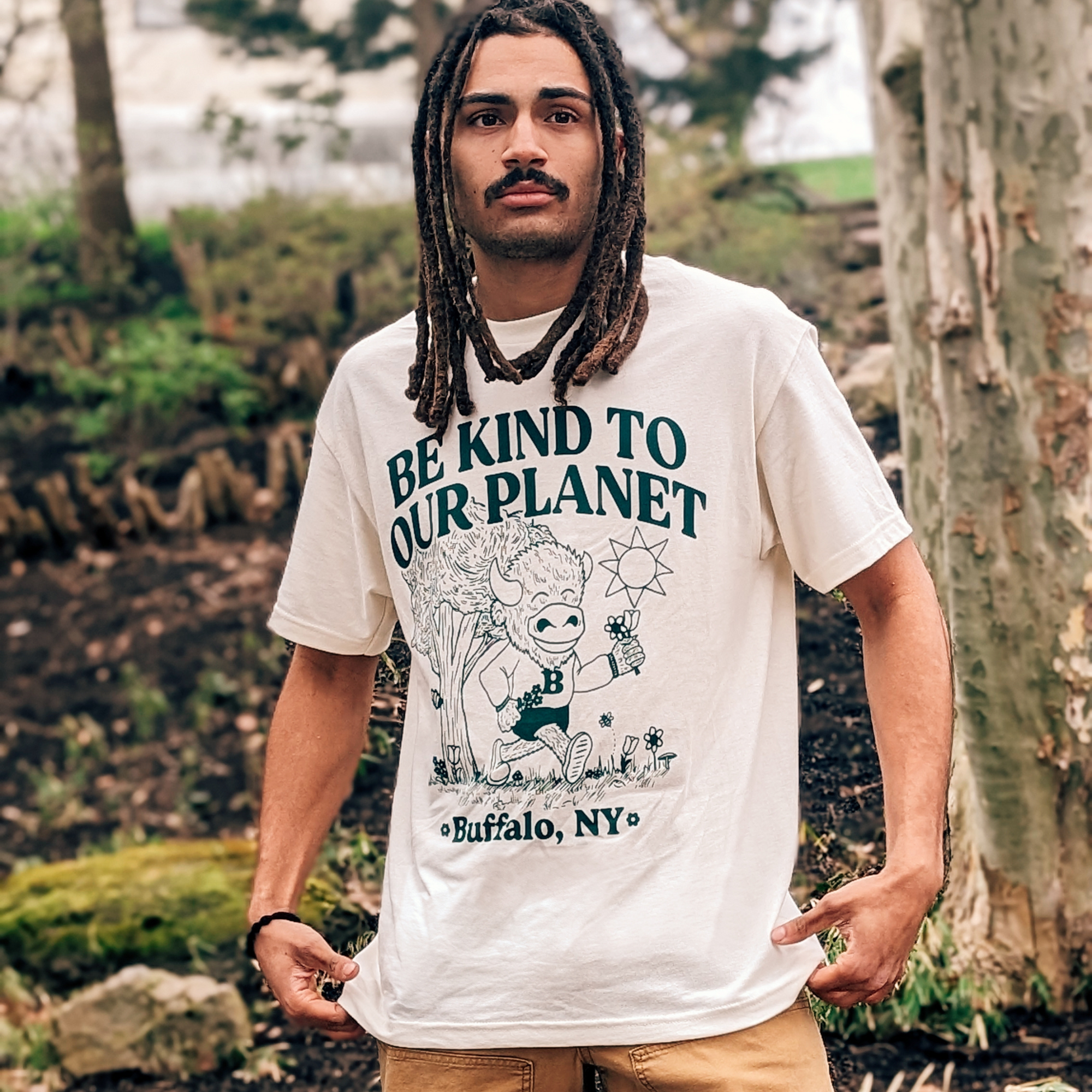 Be Kind To Our Planet Buffalo, NY Cream Short Sleeve Shirt. Shirt features a drawing of a buffalo smiling and walking through a park.
