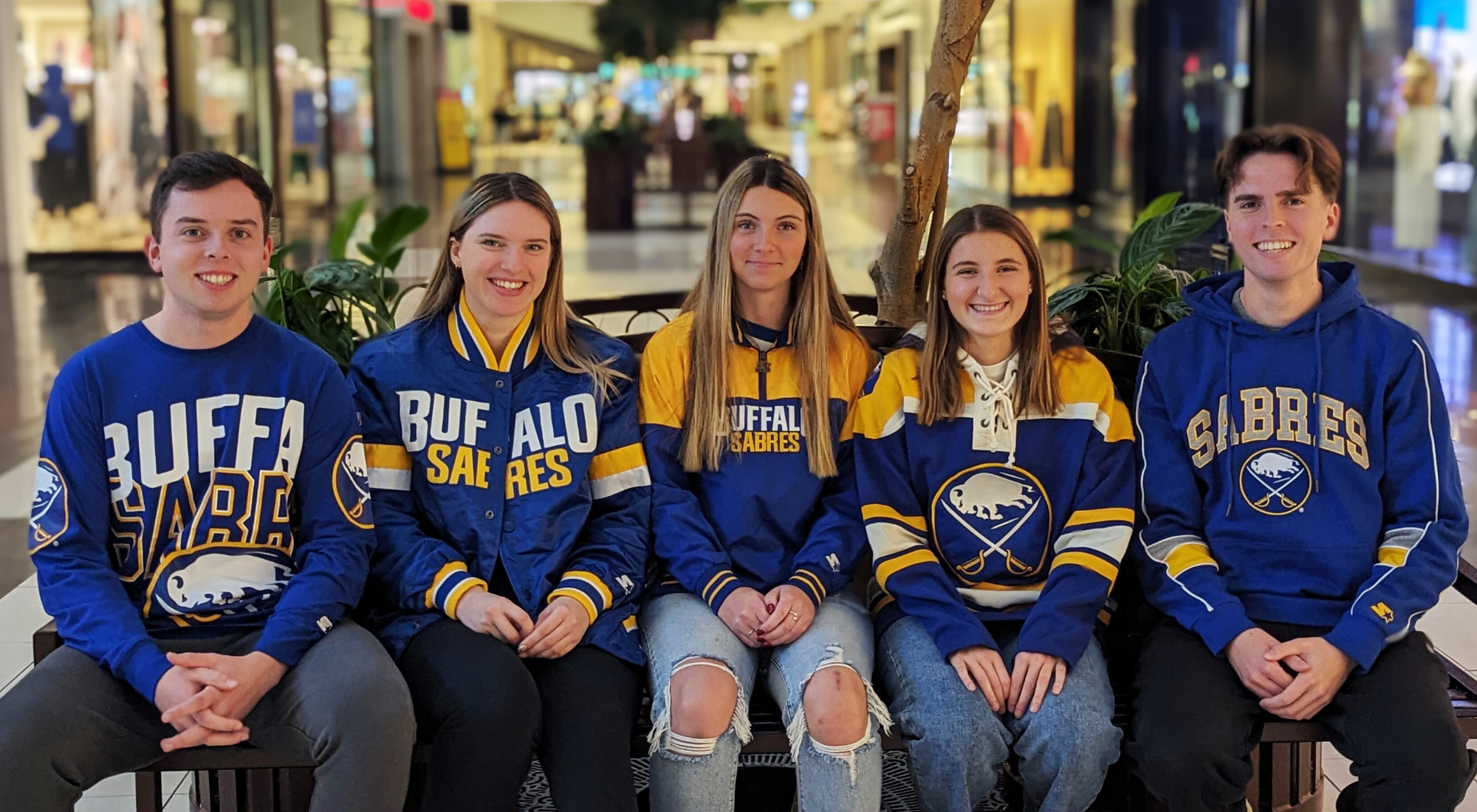 Five individuals sitting on a bench in the mall posing for a picture wearing buffalo sabres apparel