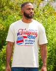 A man wearing a cream-colored graphic t-shirt with a 'Tailgate Club' Buffalo, NY design, standing outdoors in front of green foliage.