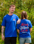 A man and a woman standing outdoors in front of green bushes, both wearing bright blue graphic T-shirts. The man's shirt has a small white and red logo on the left chest, and the woman's shirt has a large red and white graphic on the back reading 'Nobody Tailgates Like Us Buffalo, NY'.