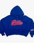 A royal blue cropped hoodie featuring 'Bills' in red cursive script with a white outline across the chest, and a Buffalo Bills logo on the left sleeve, shown against a plain light gray background.