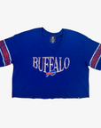 A royal blue Buffalo Bills cropped V-neck t-shirt with a distressed white 'BUFFALO' text graphic and red, white, and blue sleeve stripes, shown flat on a light gray background.