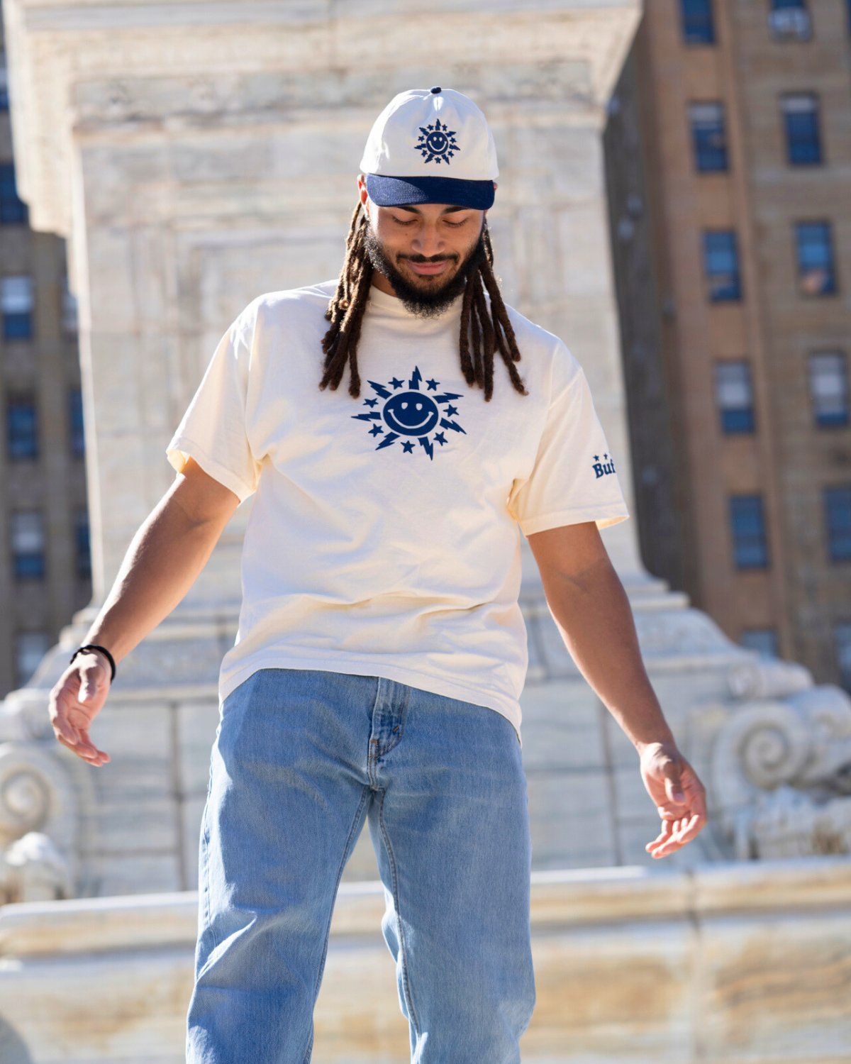 Man wearing a white t-shirt with a blue logo and a cap, standing in front of a building.