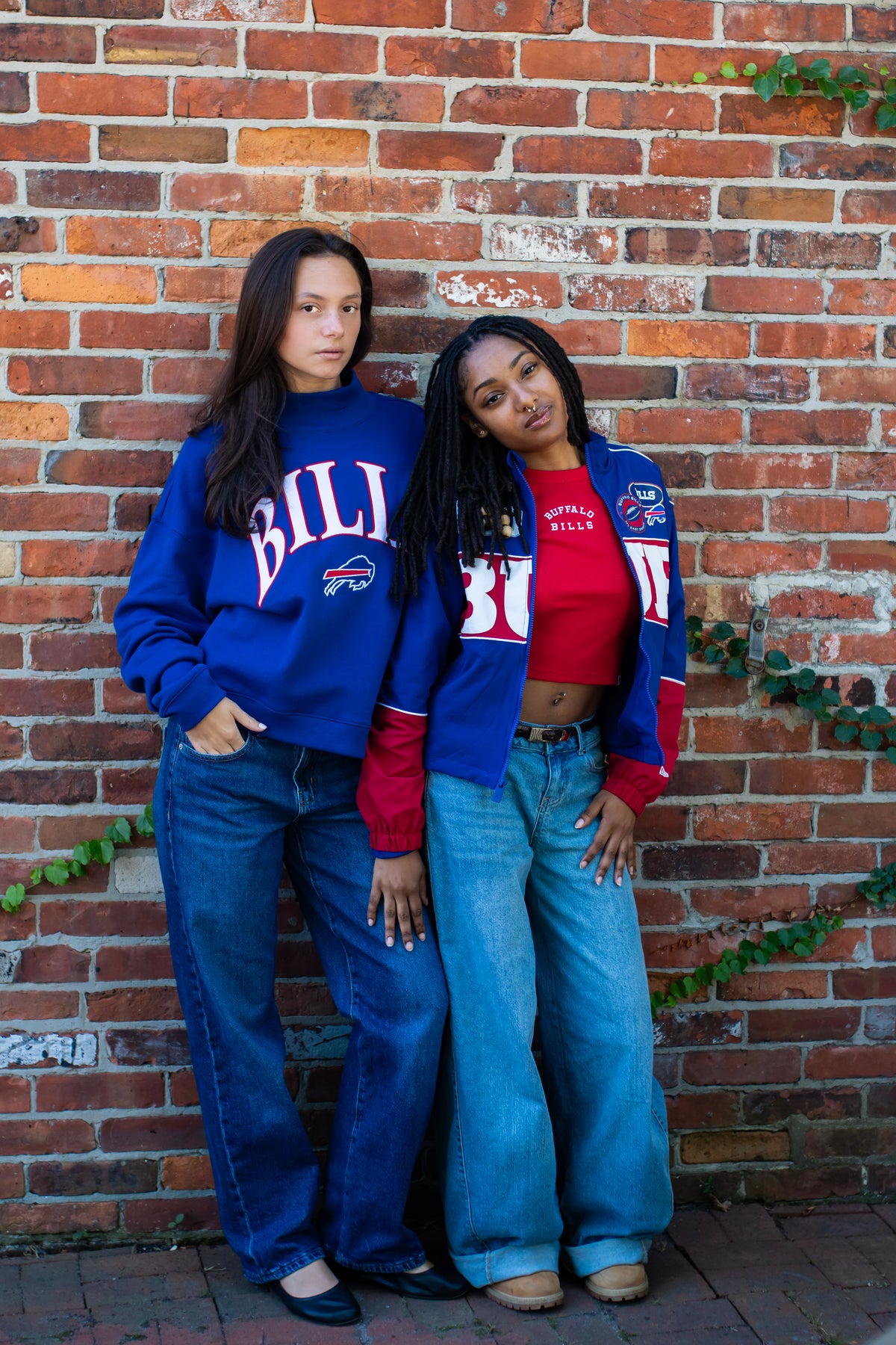 Two women posing in front of brick wall one women wearing royal Bills mock crewneck and other wearing royal blue Bills windbreaker and a red cropped tee embroidered with Buffalo Bills 