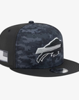 A flat-brim baseball cap featuring a dark digital camouflage pattern on the front panels and a solid black crown and brim. The front displays a white and gray Buffalo Bills charging logo, and the side has a small white American flag patch.