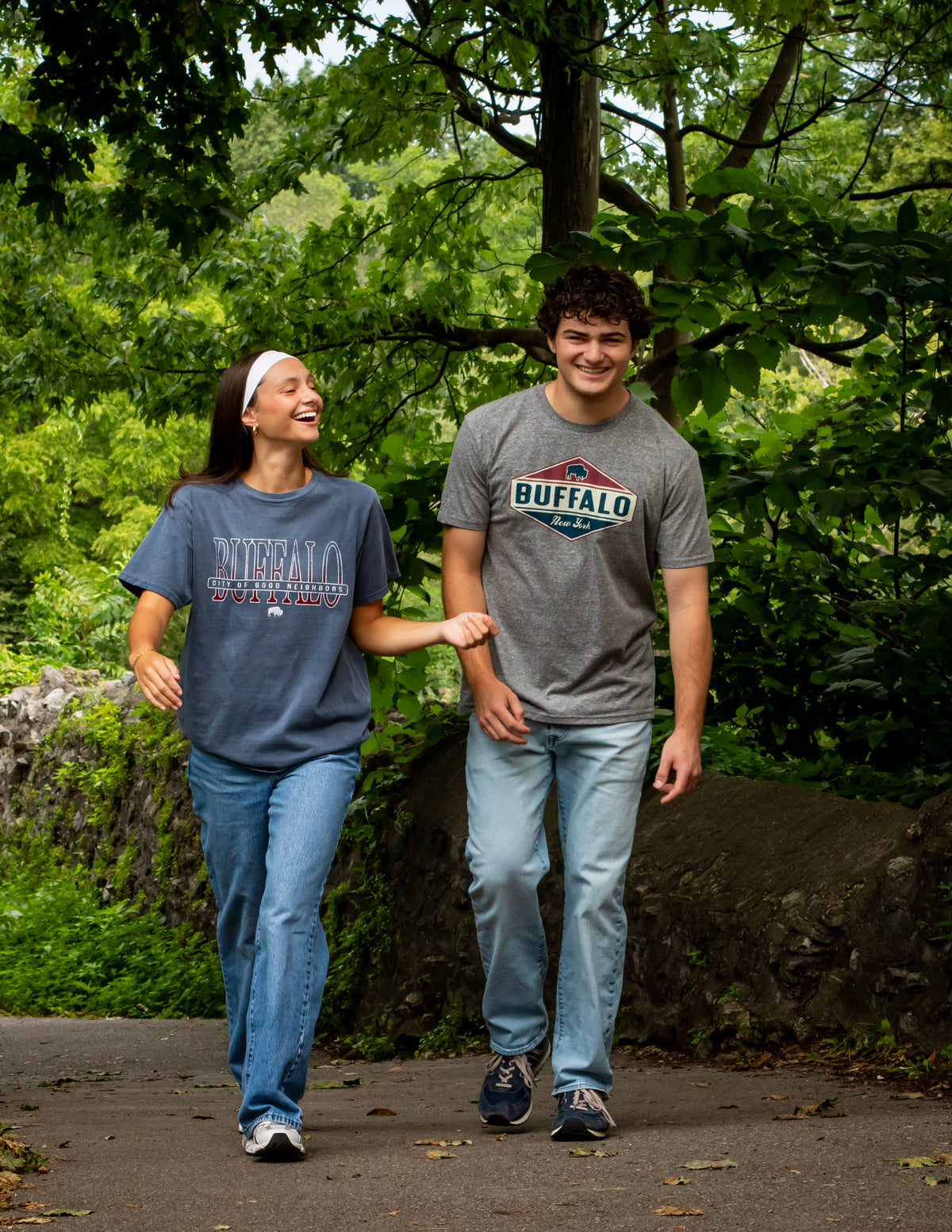 Two models, one guy and one girl, walking together laughing candidly. The woman model is wearing a washed Blue -shirt that says Buffalo in ombre blue to red, in the middle of the word Buffalo it says City of Good neighbors cutting right across.  The male model is wearing a grey t-shirt, in the middle is red, cream and Blue diamond. At the top of the diamond is a blue Buffalo logo and then it says the word Buffalo in Blue and then it says New York in cursive in cream.
