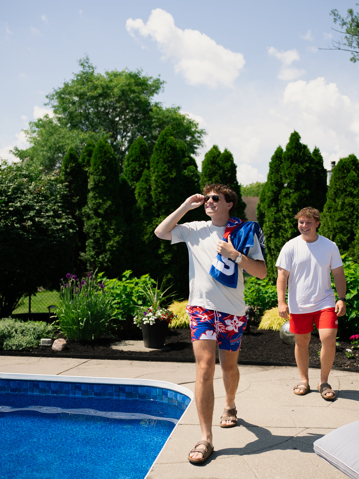 Two male models walking past a pool candidly. The first model is wearing a white t-shirt, Hawaiian floral Buffalo Bills swim trunks and carrying a royal blue buffalo bills towel. The second model is wearing a white t-shirt and red Buffalo Bills swim trunks. 