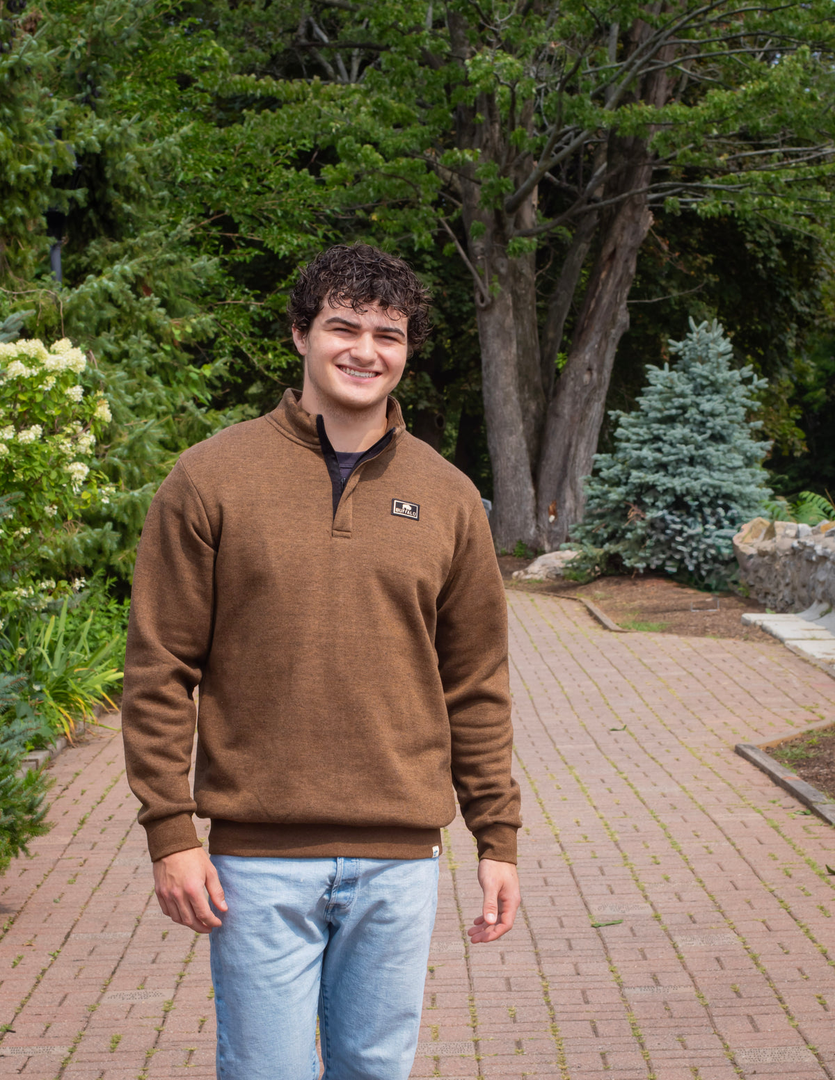 One male model, posing on a brick road with trees in the background. The model is wearing a brown quarter zip sweatshirt. On the right hand side of the chest area there is a small black and cream patch that reads Buffalo in cream and then there is a small buffalo logo in cream as well.