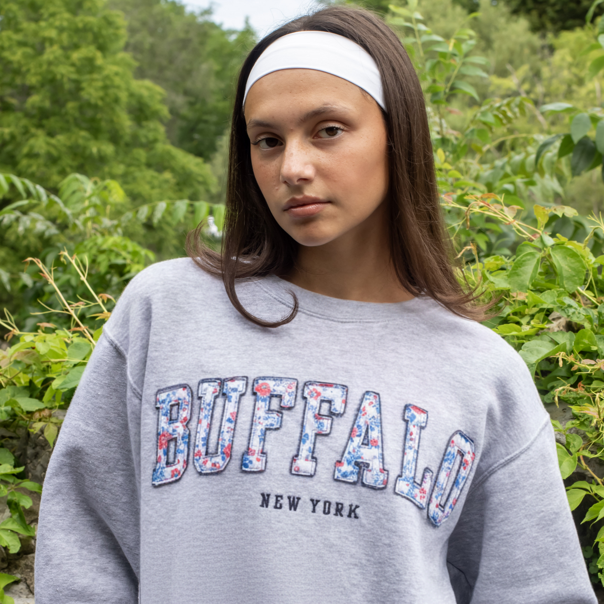 A women model wearing a grey crewneck sweatshirt that says Buffalo, NY on it, the word Buffalo is written out in floral patches stitched onto the crewneck, the flowers are red and blue.