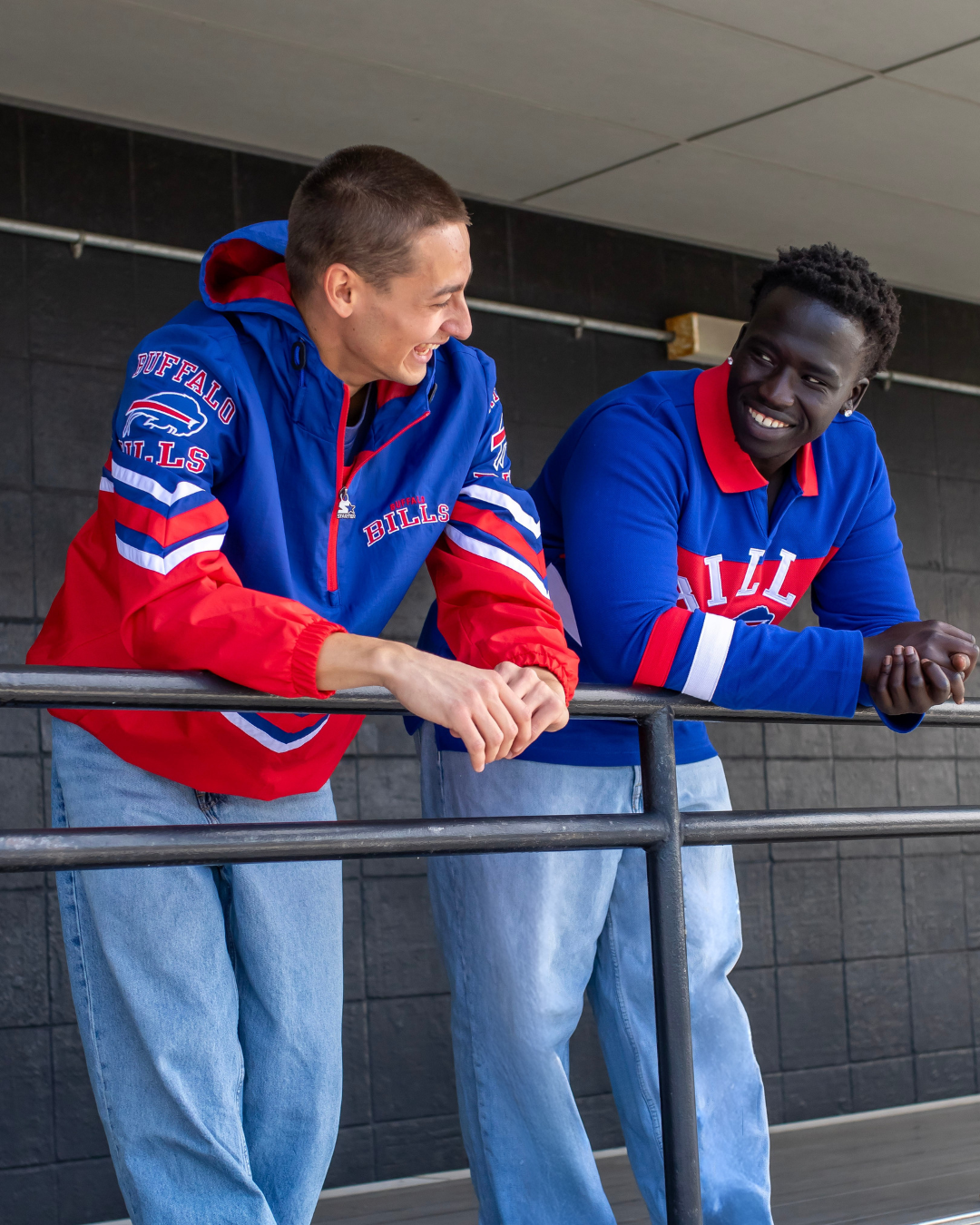 Two male models, both leaning on a railing to pose. The model to the left is wearing a royal blue and red Buffalo Bills windbreaker quarterzip jacket.  On the top right hand side of the jacket the words Buffalo Bills on it in red. On the left sleeve of the jacket is also says Buffalo Bills with a Buffalo Bills logo, The model on the right is wearing a royal blue and red polo top.  The color of the collar is red and it says Bills across the chest in white embroidery. 