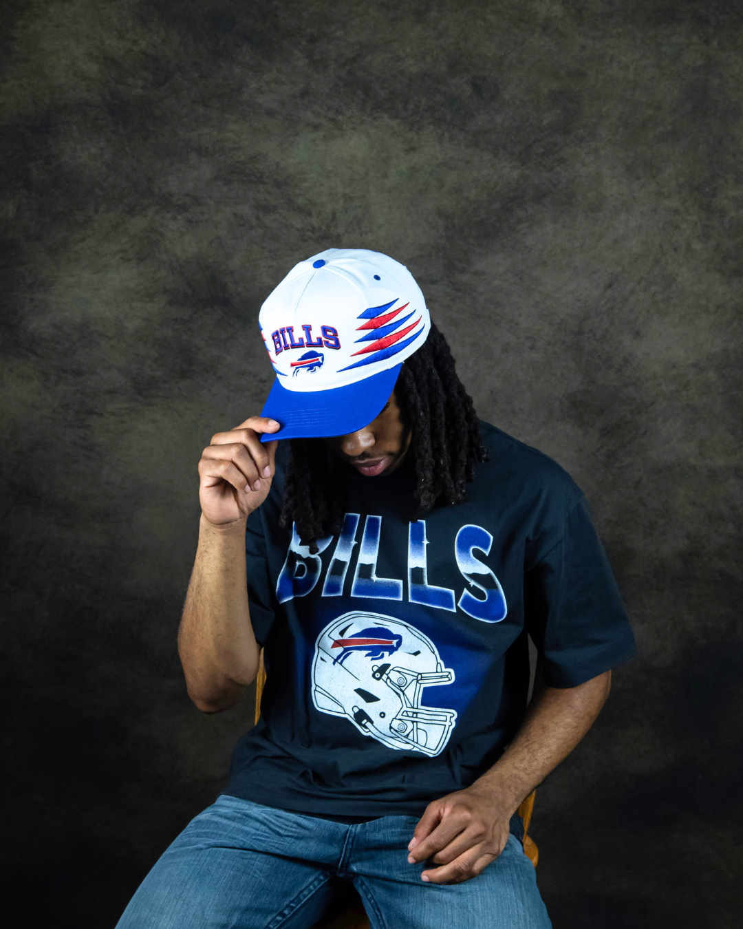 One model is posing wearing a white Bills hat. The hat has red and blue stretched out diamonds on the sides and it says Bills across the middle in royal blue with a Bills logo underneath. The model is also wearing a Black Bills t-shirt that says Bills in a shiny Blue/silver color and then there is a white football helmet underneath.