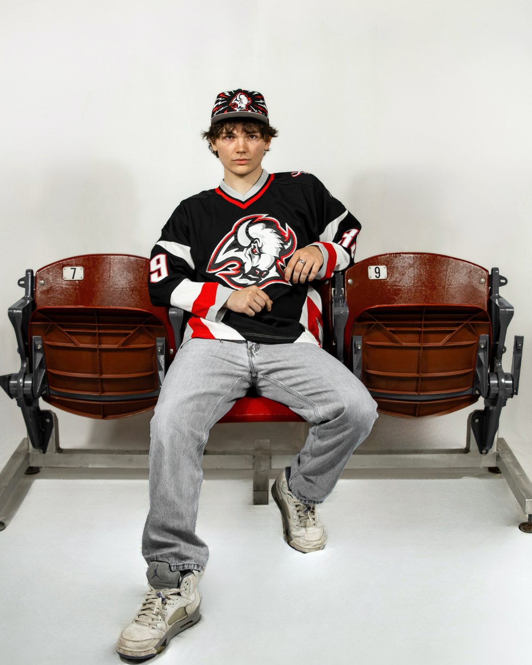 One model is sitting on old stadium chairs, wearing a red and black Sabres jersey with the goat-head Sabres logo on it. He is also wearing a black Buffalo Sabres cap with the goat head logo in the middle. Around the hat are lightning bolts in red and white.