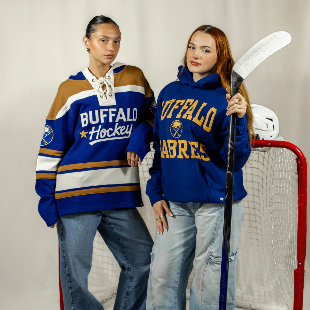 Two women posing, one is wearing a Blue hoodie that says Buffalo Sabres on it, the other is wearing a sweatshirt that ties by the neckline and says Buffalo Hockey on it with a yellow star embroidered patch next to it.