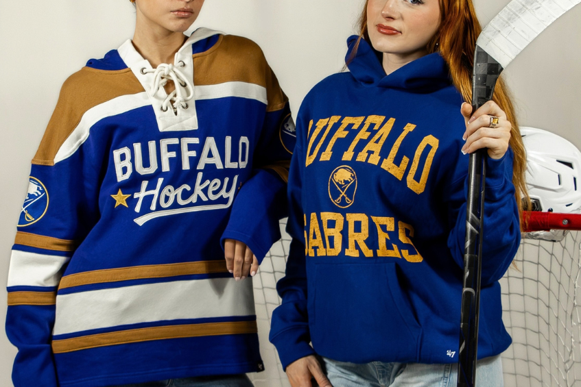 Two women posing in front of a hockey net and one is holding a hockey stick.  The model on the left is wearing a hoodie that is mocking a hockey jersey, it has a tie up design at the neckline with white strings, The stripes going across the sweatshirt are mustard yellow and royal blue and it says Buffalo hockey with a yellow star. The model on the right is wearing a royal blue hoodie that says Buffalo Sabres in yellow.