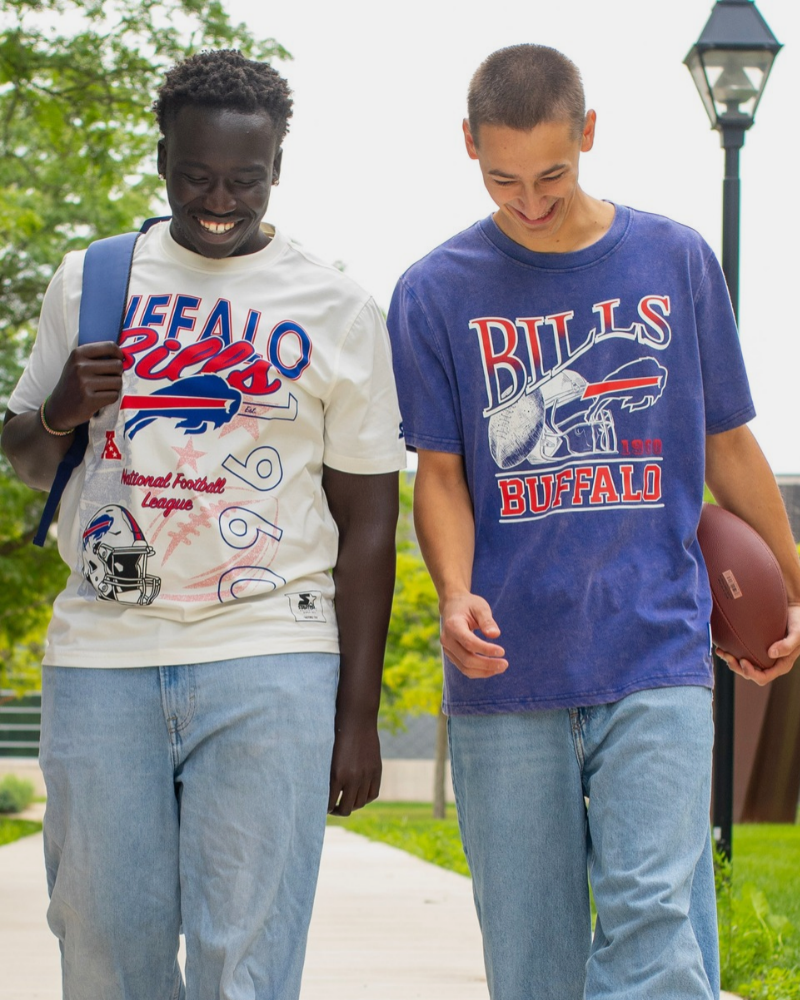 Two men wearing vintage Buffalo Bills graphic t-shirts and light-wash jeans, walking outdoors with a football.