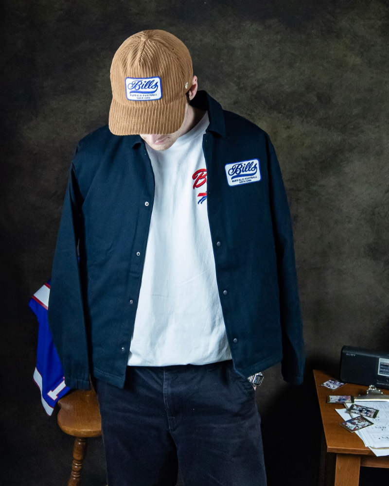 Model wearing a black Buffalo Bills jacket over a white graphic t-shirt, paired with a tan corduroy Bills hat and dark jeans.