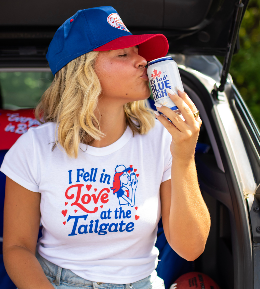 A woman wearing a white t-shirt with a red and blue 'I Fell in Love at the Tailgate' graphic, a blue and red baseball cap, and holding a can of Blue light while leaning against the open trunk of a car.