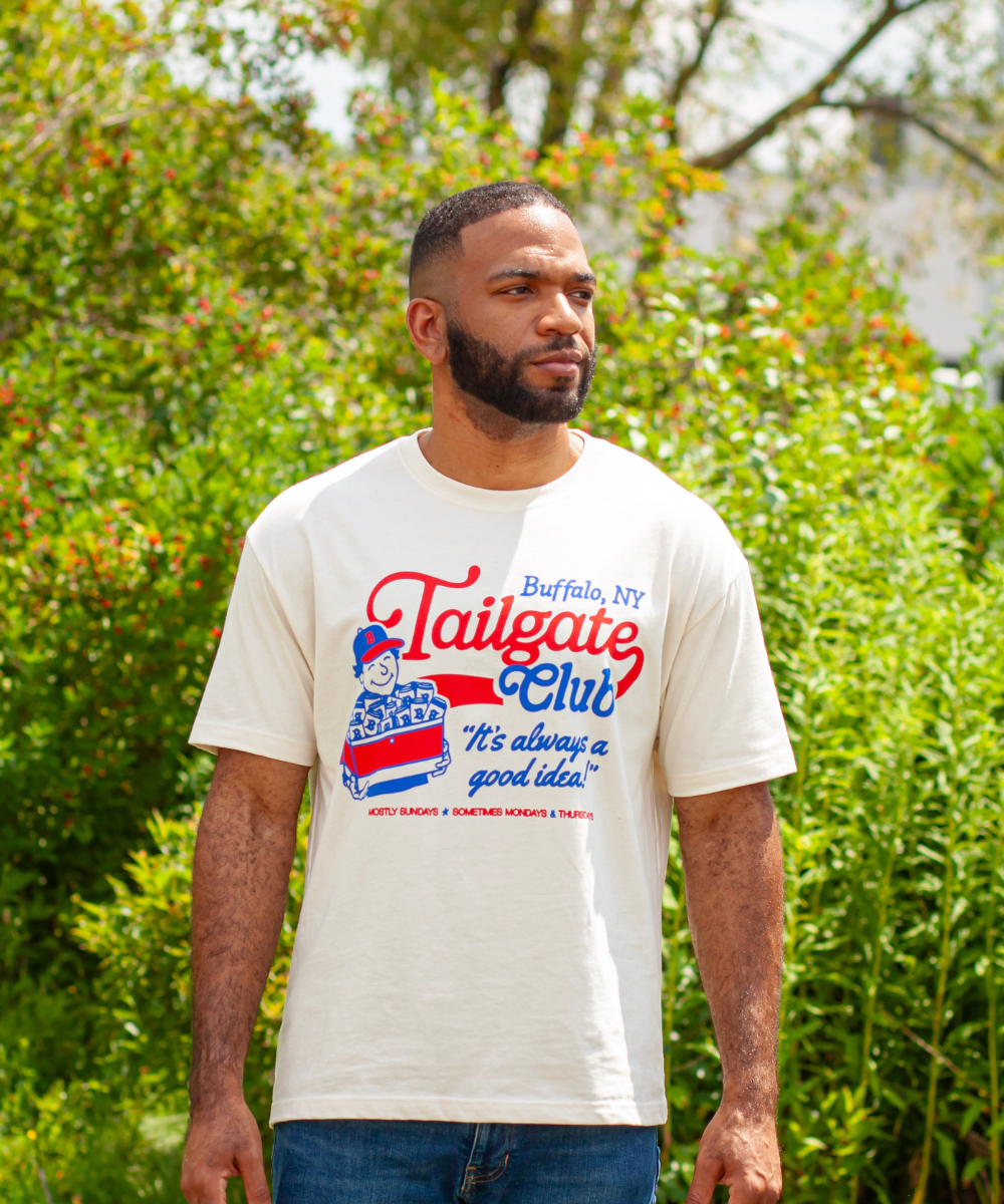 A man wearing a white graphic t-shirt with a red and blue 'Tailgate Club' Buffalo, NY design, standing outdoors in front of green foliage.