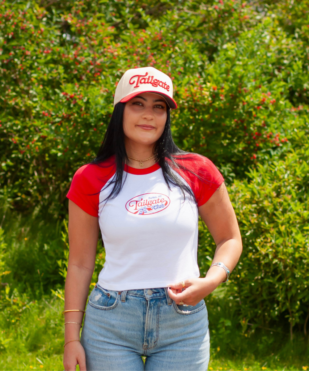 A woman wearing a red and white raglan t-shirt with a 'Tailgate' logo, paired with a matching baseball cap and light blue jeans, standing outdoors in front of a green hedge.