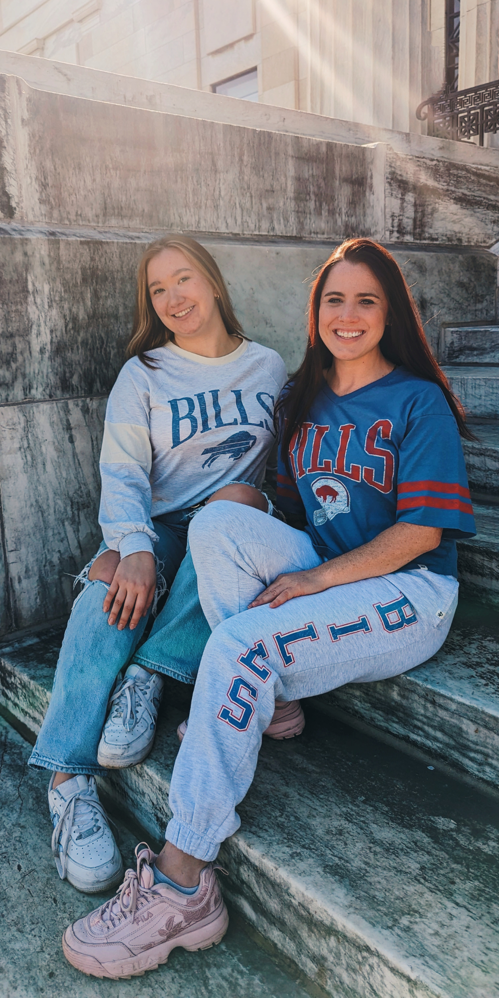 two woman posing for a picture one wearing buffalo bills blue short sleeve shirt and gray bills sweatpants and the other lady wearing a gray bills long sleeve shirt