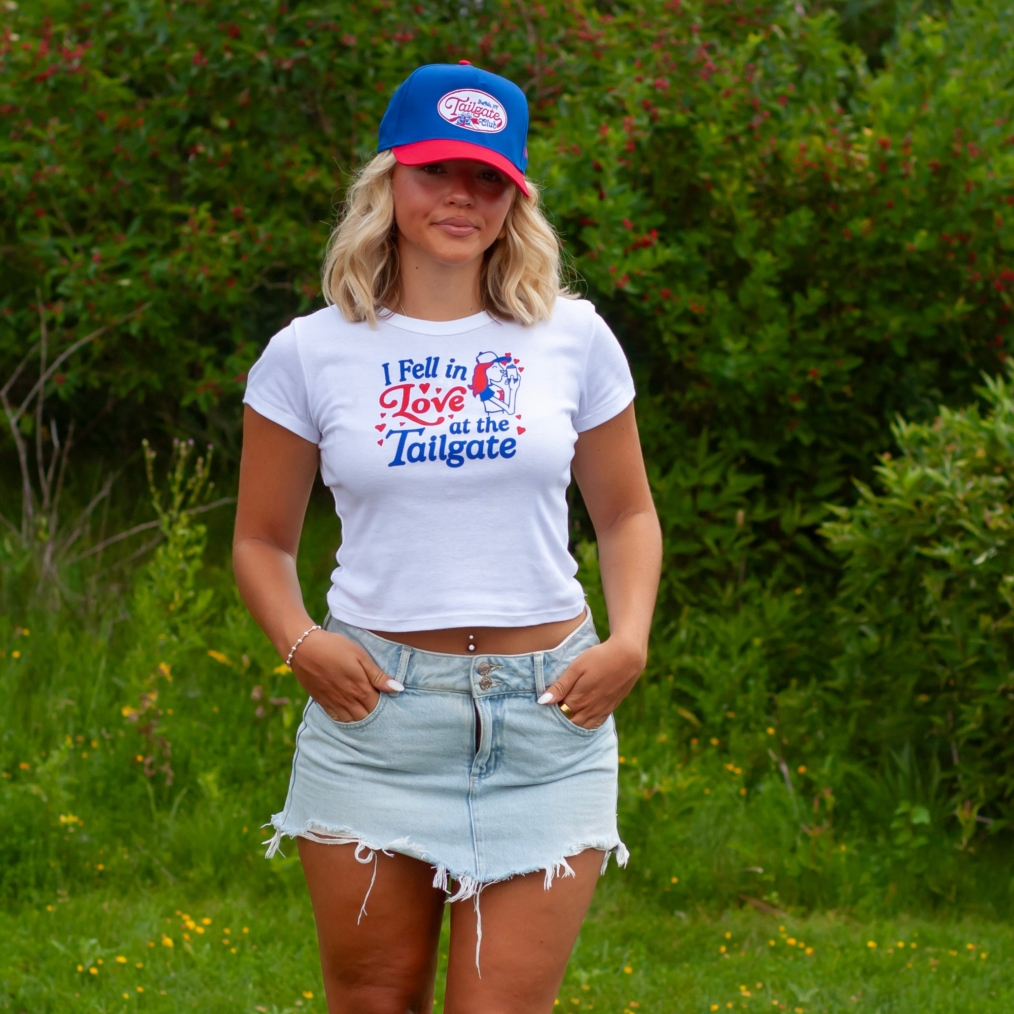 A woman standing outdoors in front of green bushes, wearing a white graphic t-shirt with a red and blue 'I Fell in Love at the Tailgate' design, a blue and red trucker hat, and a light-wash denim skirt.