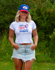 A woman standing outdoors in front of green bushes, wearing a white graphic t-shirt with a red and blue 'I Fell in Love at the Tailgate' design, a blue and red trucker hat, and a light-wash denim skirt.