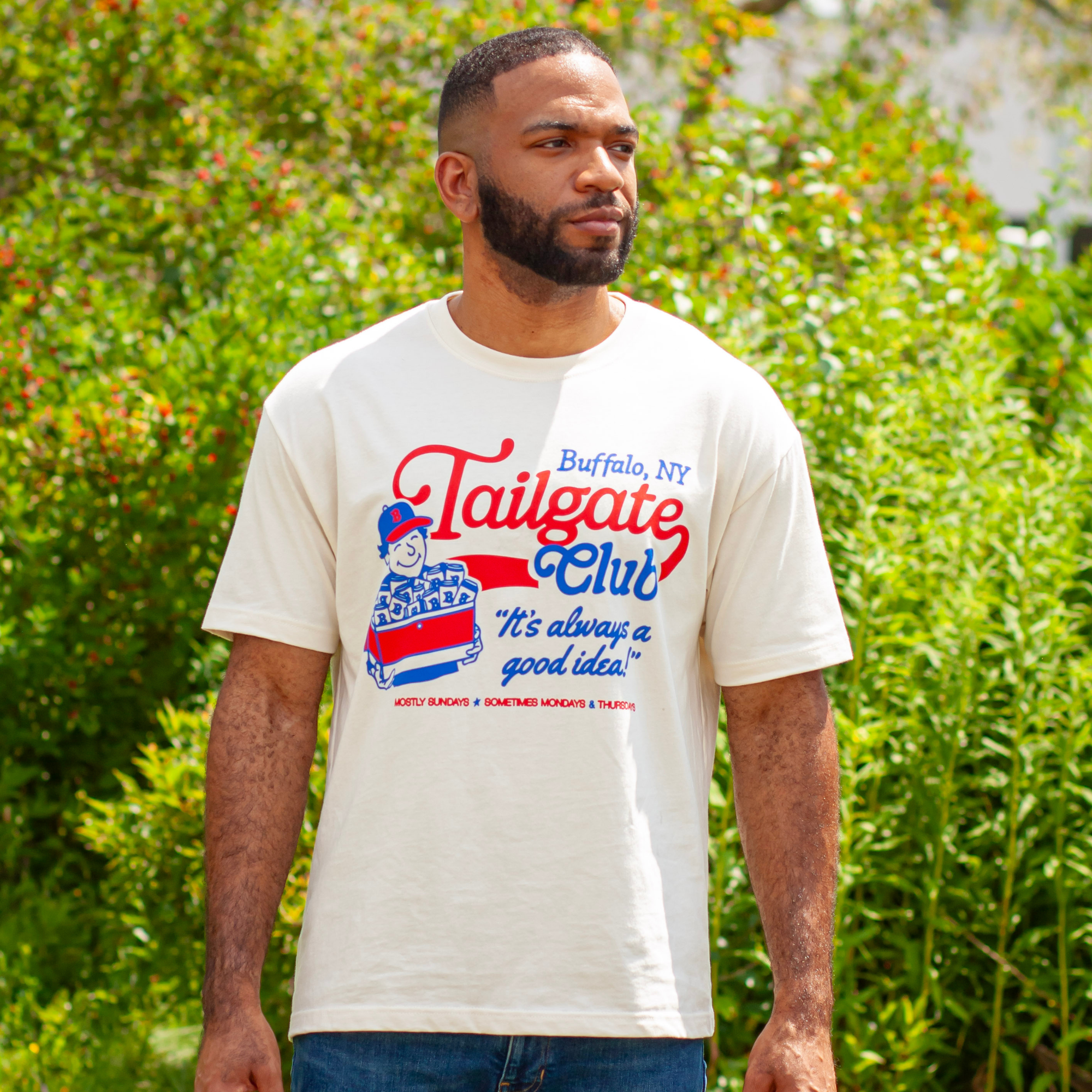 A man wearing a cream-colored graphic t-shirt with a 'Tailgate Club' Buffalo, NY design, standing outdoors in front of green foliage.
