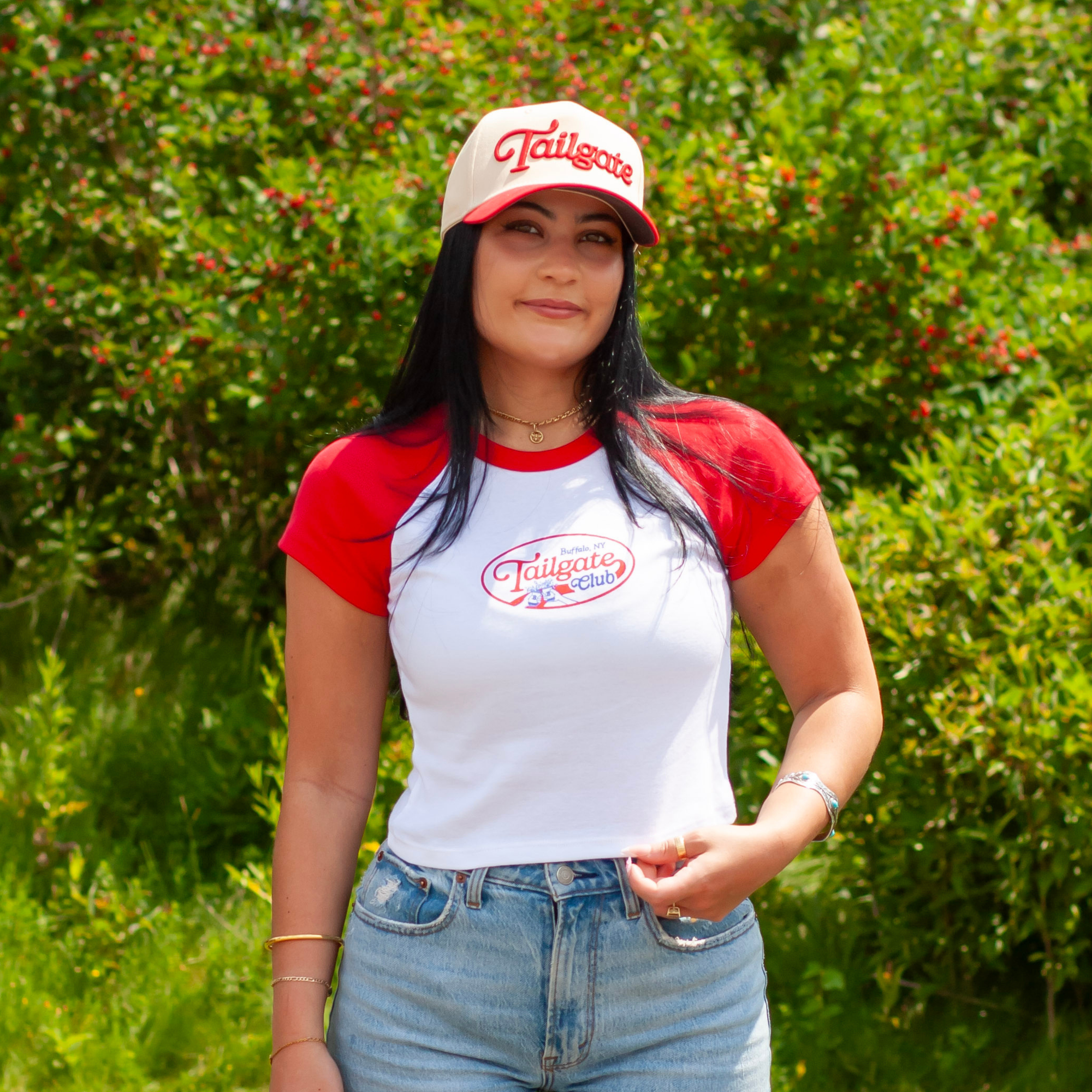 A woman wearing a white and red raglan t-shirt with a 'Tailgate with Us' logo, matching baseball cap, and blue jeans, standing outdoors in front of a green hedge.