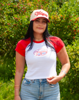 A woman wearing a white and red raglan t-shirt with a 'Tailgate with Us' logo, matching baseball cap, and blue jeans, standing outdoors in front of a green hedge.