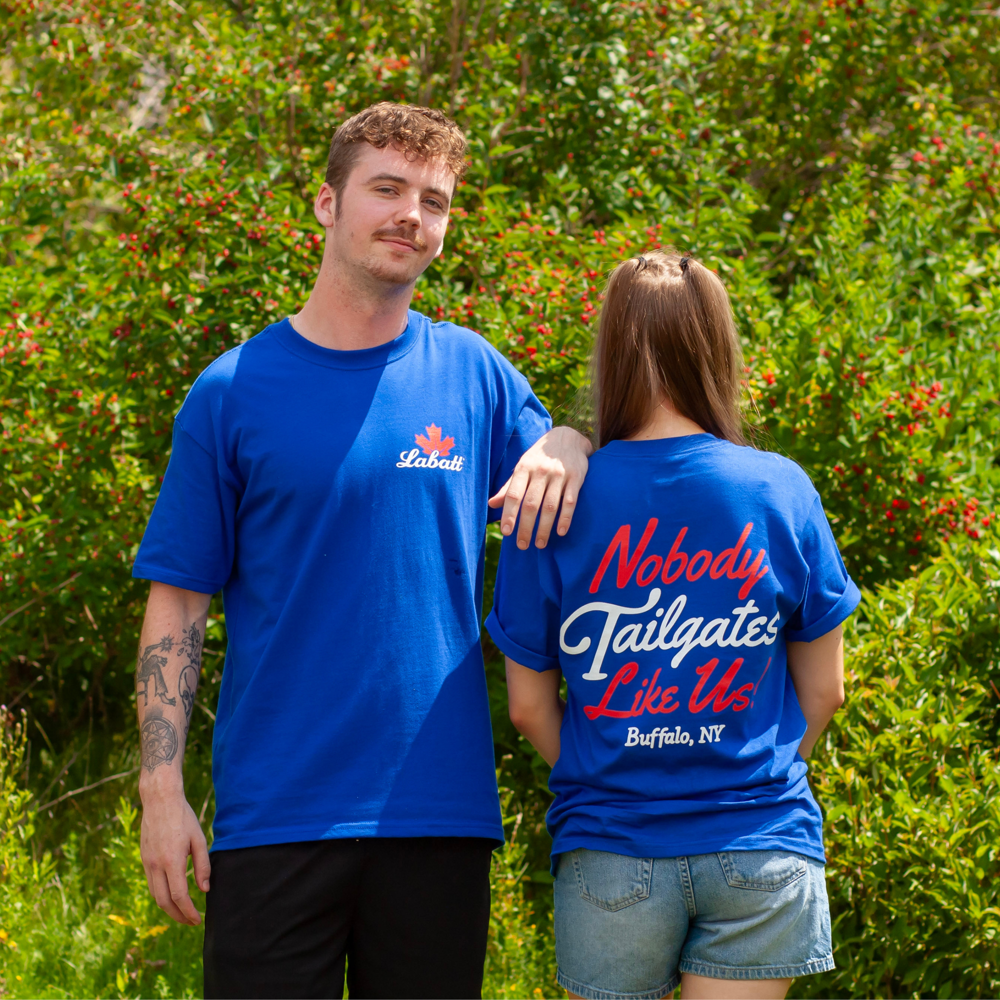 A man and a woman standing outdoors in front of green bushes, both wearing bright blue graphic T-shirts. The man's shirt has a small white and red logo on the left chest, and the woman's shirt has a large red and white graphic on the back reading 'Nobody Tailgates Like Us Buffalo, NY'.