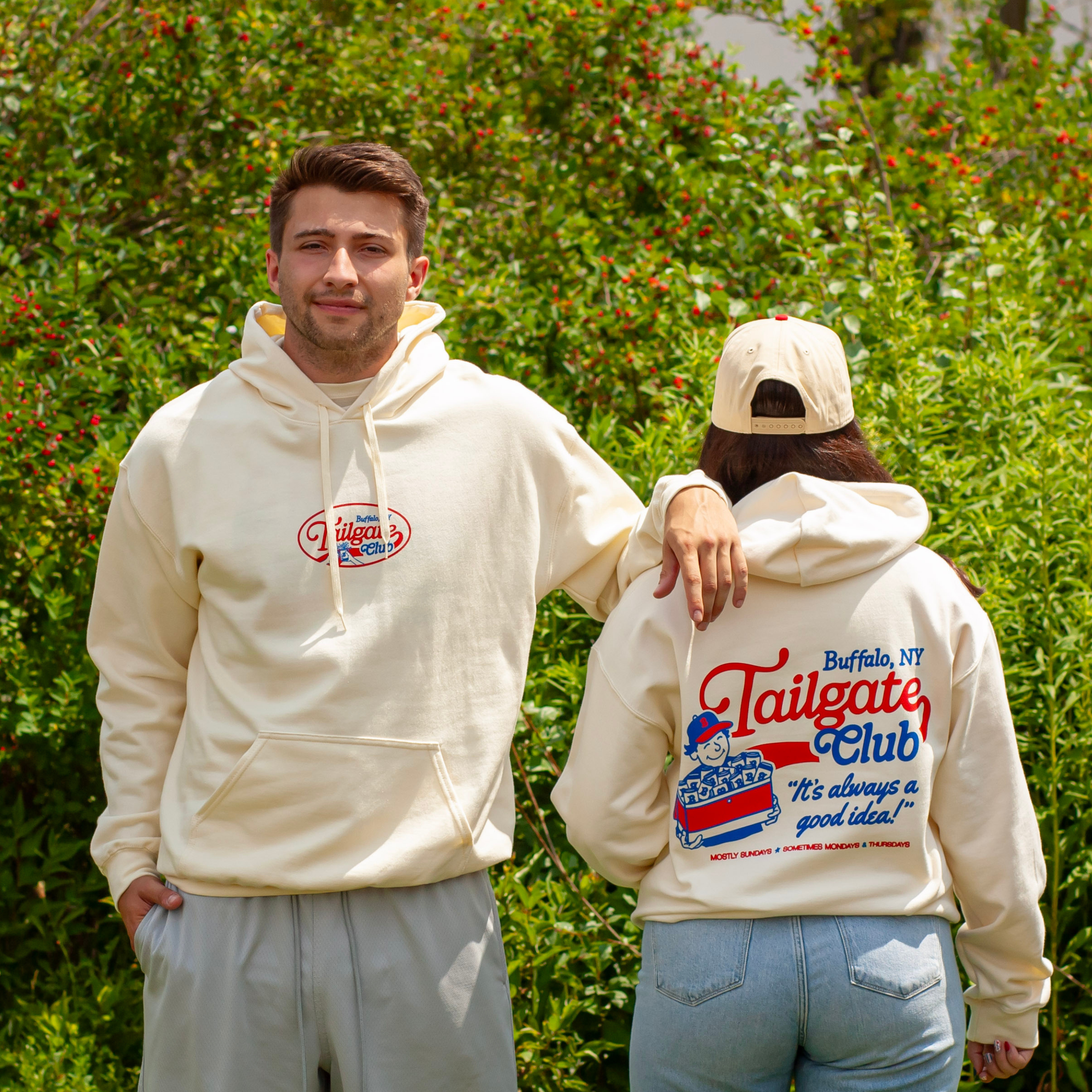 A man and a woman wearing matching cream-colored hoodies with Buffalo, NY Tailgate Club graphics, standing outdoors in front of green foliage.