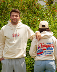 A man and a woman wearing matching cream-colored hoodies with Buffalo, NY Tailgate Club graphics, standing outdoors in front of green foliage.
